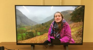 A large television screen, showing Katie sitting on a misty mountainside, speaking to the camera