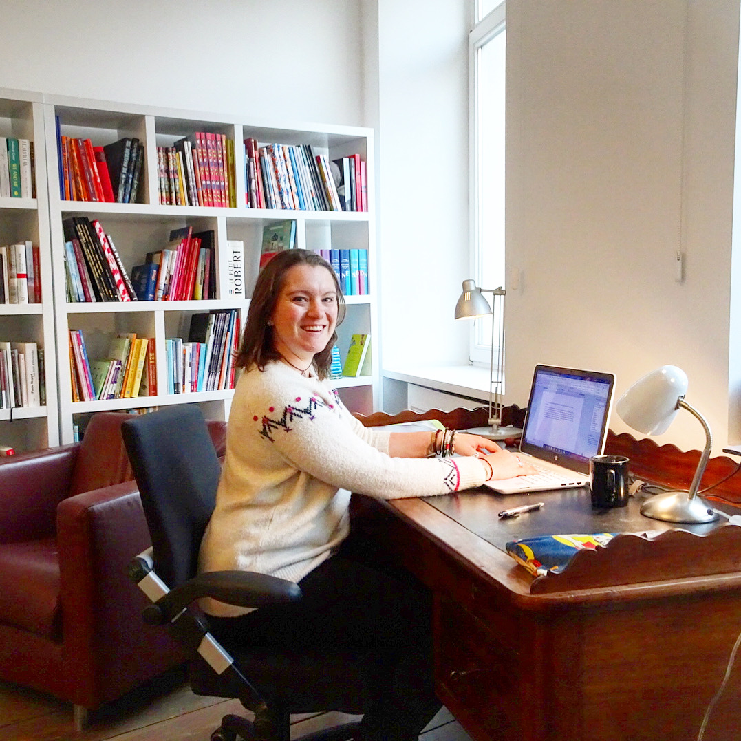Katie sitting at a desk in a light airy room in a Brussels apartment, with shelves of books behind her