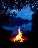 Nighttime; lake and mountains through a gap in the trees; on a stony beach, a brazier with a campfire burning