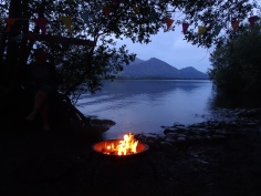 Nighttime; lake and mountains through a gap in the trees; on a stony beach, a brazier with a campfire burning
