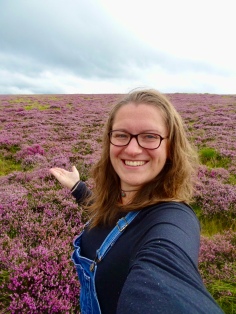 Katie wearing glasses and dungarees and smiling at the camera; behind, purple heather stretching away across the fell