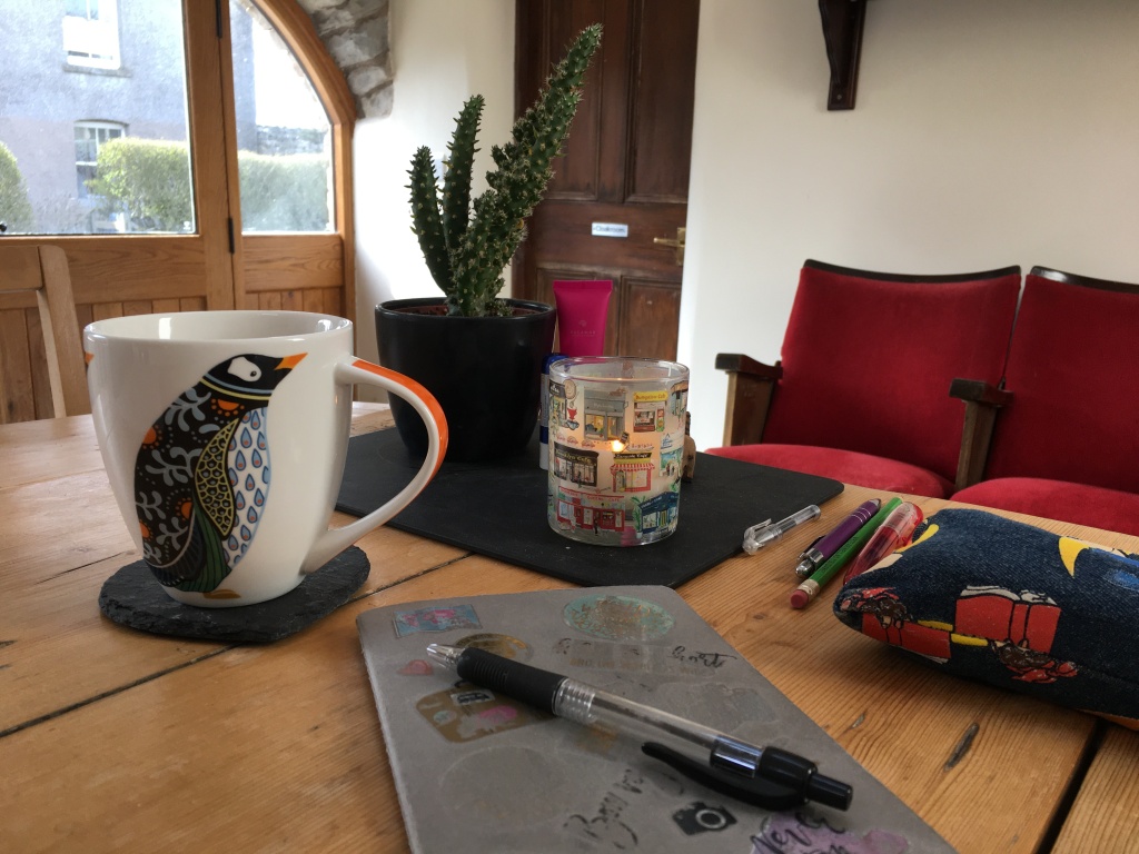 A kitchen table, with a cactus, a lit candle, a notebook and pen, and a mug of coffee