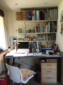 A desk covered in bits of manuscript, with shelves of notebooks above