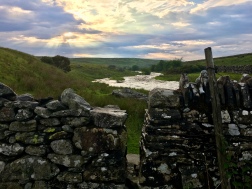 A dry stone wall with a stile in it. Beyond it, the River Lowther, and a cloudy sky above, with god rays shining through