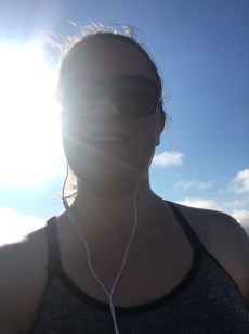 Katie with sunglasses and earbuds, smiling down at the camera with a blue sky and the sun behind her