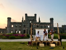 Three actors playing musical instruments on a temporary stage, with a festoon of lights above it; a silhouetted castle and a sunset in the background